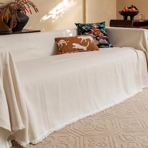 May include: A white throw blanket with a fringed edge draped over a white couch. The couch has two pillows, one with a colorful geometric pattern and the other with a brown and white animal print.