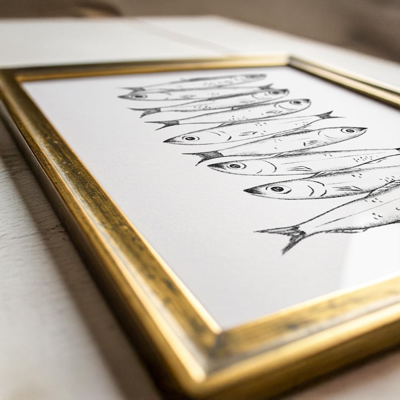 May include: A framed art print featuring a series of stylized fish illustrations in black ink on white paper. The frame is a gold color, adding a touch of elegance to the artwork. The print is angled, showing the depth of the frame.