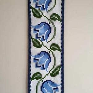 May include: A handcrafted wall hanging with a floral pattern. The rectangular piece displays blue flowers and green leaves against a white backdrop, enclosed by a dark blue border. It is hung from a wooden dowel with a white cord.