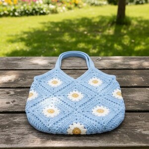 Puede incluir: Un bolso tote de ganchillo azul claro con un estampado de margaritas. El bolso presenta aplicaciones de margaritas blancas y amarillas sobre un patr&oacute;n cuadrado. El bolso tiene dos asas azules claras y est&aacute; sobre un banco de madera.