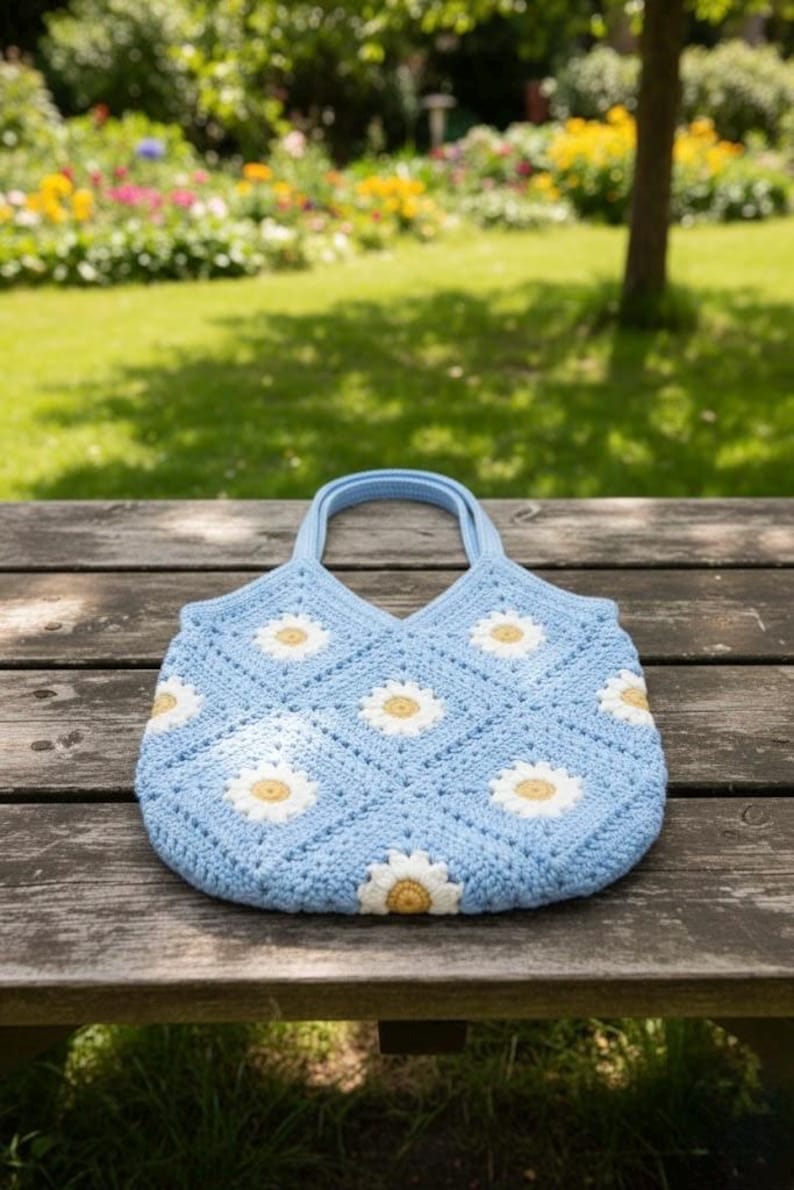 Puede incluir: Un bolso tote de ganchillo azul claro con un estampado de margaritas. El bolso presenta aplicaciones de margaritas blancas y amarillas sobre un patr&oacute;n cuadrado. El bolso tiene dos asas azules claras y est&aacute; sobre un banco de madera.