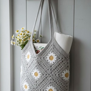 Può includere: Una borsa tote grigia all'uncinetto con applicazioni di margherite bianche e gialle è appesa a un gancio di legno. La borsa è riempita con un mazzo di margherite, un libro e una coperta bianca arrotolata. La borsa è sospesa contro un muro grigio.