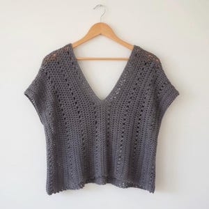Puede incluir: Un top de ganchillo gris, de manga corta y cuello en V profundo. El top está colgado de una percha de madera sobre un fondo blanco. El patrón de ganchillo presenta detalles calados.