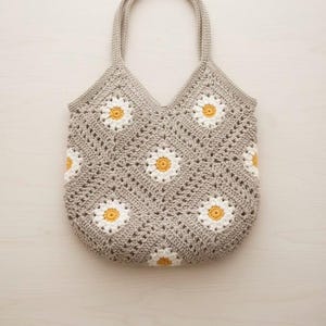 Puede incluir: Un bolso tote tejido a ganchillo en color gris claro, con un patr&oacute;n de margaritas con p&eacute;talos blancos y centros amarillos. El bolso tiene dos asas y una parte superior en forma de V. El patr&oacute;n de margaritas se repite en la parte delantera y trasera del bolso.