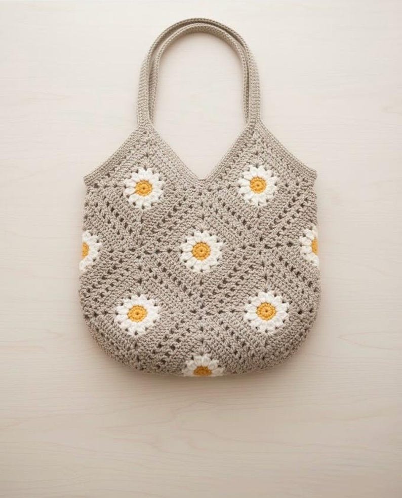Puede incluir: Un bolso tote tejido a ganchillo en color gris claro, con un patr&oacute;n de margaritas con p&eacute;talos blancos y centros amarillos. El bolso tiene dos asas y una parte superior en forma de V. El patr&oacute;n de margaritas se repite en la parte delantera y trasera del bolso.