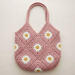Puede incluir: Un bolso tote rosa tejido a crochet con un estampado de margaritas. El bolso tiene una parte superior en forma de V y dos asas. El estampado de margaritas presenta p&eacute;talos blancos y centros amarillos, creando un dise&ntilde;o floral encantador.