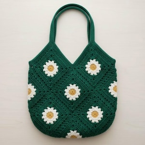 Puede incluir: Bolso tote de crochet verde hecho a mano con aplicaciones de margaritas blancas y amarillas. El bolso tiene dos asas verdes y una parte superior en forma de V. El patr&oacute;n de margaritas se repite en la parte delantera y trasera.