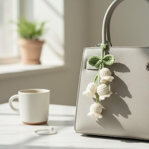 May include: A grey leather handbag with a crocheted lily of the valley charm. The charm features white flowers and green leaves. A white mug and a silver bracelet are on the table.
