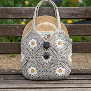 Puede incluir: Bolso tote de crochet gris con detalles de margaritas blancas y amarillas. El bolso contiene un sombrero de paja y gafas de sol. Descansa sobre un banco de madera, con un fondo borroso de flores y follaje.