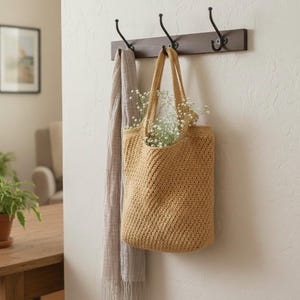 Può includere: Una borsa tote all'uncinetto color beige piena di fiori bianchi è appesa a un attaccapanni in legno marrone scuro con tre ganci in metallo nero. Una sciarpa grigio chiaro è appesa a uno dei ganci. Il muro è dipinto di beige chiaro.