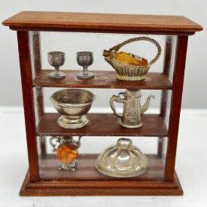 May include: A wooden display cabinet with glass doors and three shelves. The shelves are filled with miniature silver and glass items, including a wine bottle, glasses, a bowl, a teapot, and a covered dish.