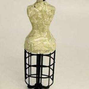 May include: A miniature dress form mannequin featuring a cream-colored torso with a subtle, light-toned pattern. The torso is supported by a black metal cage-style skirt frame and a black tripod base, topped with a small black finial knob.