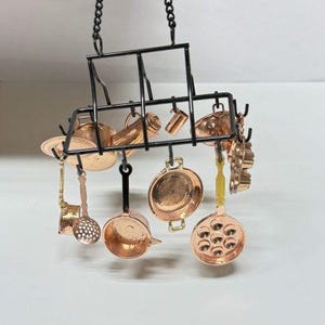 May include: Miniature copper kitchen cookware set hanging from a black metal rack. The set includes pots, pans, a ladle, and other cooking utensils. The rack is suspended by a chain.
