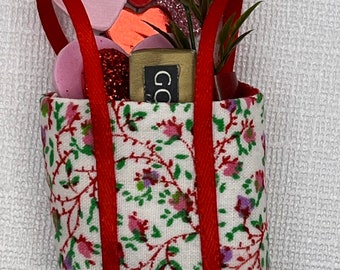 May include: A fabric tote bag with a floral pattern, red handles, and various heart-shaped decorations. The bag contains a small box with the letters "GO" and sprigs of greenery. The hearts are in shades of red and pink.