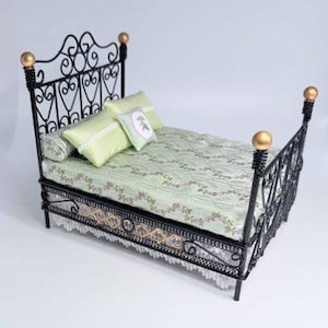 May include: A miniature dollhouse bed featuring a black wrought iron frame with gold-toned finials. The bed is dressed in a light green patterned mattress cover, a matching bolster pillow, a rectangular pillow, and a small square floral pillow.