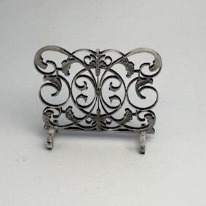 May include: A silver-colored, decorative metal screen with an ornate, symmetrical design. The screen features swirling patterns and a central floral motif. It stands on two small feet, and is likely a miniature decorative piece.