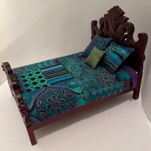 May include: Miniature wooden bed with a carved headboard and footboard. The bed is covered with a patchwork quilt in shades of teal, blue, and purple. Two patterned pillows and a small green pillow complete the bedding. A charming dollhouse furniture piece.