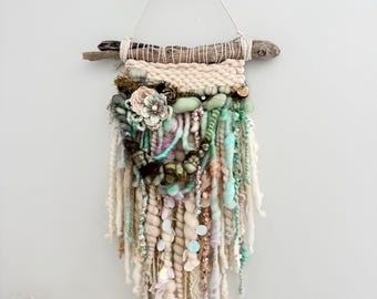 May include: A handcrafted fiber art wall hanging. The piece features a driftwood base with woven cream-colored yarn and cascading strands of various textures and colors, including cream, green, and pink. Embellishments include sequins and a fabric flower.