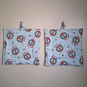 May include: Two square, light blue pot holders with a repeating pattern of Christmas wreaths, snowflakes, and candy canes. The center of each wreath has a red hand with the text "HOWDY HO!". Each pot holder has a small fabric loop.
