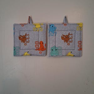 May include: Two gray, square potholders with a loop for hanging. Each features a repeating pattern of cartoon characters, including Eevee, Charmander, and Pikachu, along with numbers. The potholders are quilted with a light gray thread.