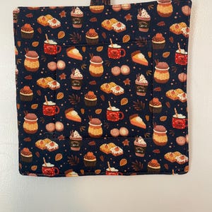May include: A black pot holder with a fall-themed print featuring cupcakes, pie, and mugs of hot drinks. The print is in shades of orange, brown, and red on a dark blue background.