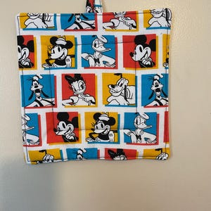 May include: Square pot holder featuring a colorful pattern of Disney characters, including Mickey Mouse, Minnie Mouse, Donald Duck, and Pluto. The design is set against a backdrop of red, yellow, and blue squares, with a blue border and a hanging loop.