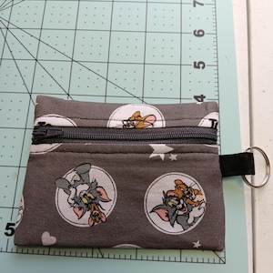 May include: A small gray zippered pouch with a Tom and Jerry cartoon print. The pouch has a black zipper and a black strap with a metal ring.