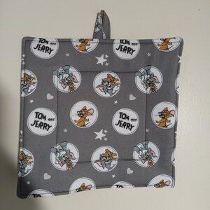 May include: A gray and white cotton pot holder with a repeating pattern of Tom and Jerry cartoon characters in circles. The text "Tom and Jerry" appears in black lettering on the pot holder.