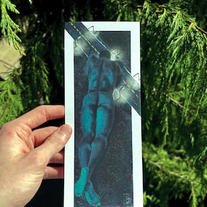 May include: An art print showing a teal figure lying down on a dark background with a road design. The figure's back and legs are visible, with teal feet. The print is held against a backdrop of green foliage.