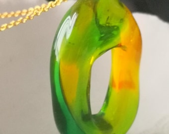 Puede incluir: Un colgante de vidrio verde y amarillo con una cadena dorada. El colgante tiene forma de anillo con un agujero en el centro.