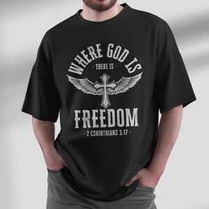 Può includere: Maglietta nera con un design grafico bianco che presenta una croce con ali e il testo "Where God is - There is - Freedom - 2 Corinthians 3:17 -"