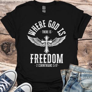Könnte beinhalten: Schwarzes T-Shirt mit einem weißen Grafikdesign, auf dem "Where God Is - There Is - Freedom - 2 Corinthians 3:17 -" mit einem weißen Kreuz und Flügeln steht.