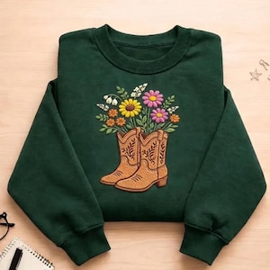 Puede incluir: Una sudadera verde oscuro con un diseño bordado de botas de vaquero marrones con un ramo de flores coloridas. Las flores incluyen flores amarillas, rosas y blancas, creando un contraste con la tela verde.