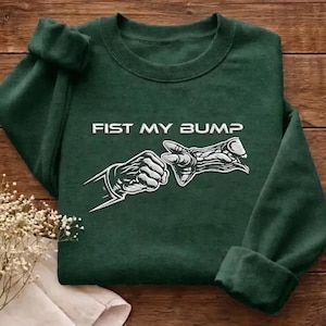 Puede incluir: Sudadera verde oscuro con el texto "FIST MY BUMP" sobre un gráfico blanco de dos manos chocando los puños. La sudadera está doblada sobre una superficie de madera.
