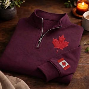 Może przedstawiać: Bordowa bluza z zamkiem 1/4, z czerwoną haftowaną liściem klonu na piersi i naszywką z flagą Kanady na rękawie. Bluza ma srebrny zamek błyskawiczny.
