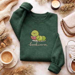May include: A dark green sweatshirt with the word "bookworm" embroidered in cursive. The sweatshirt features a cartoon caterpillar reading a book. The image includes a cup of coffee, a candle, and a purse.