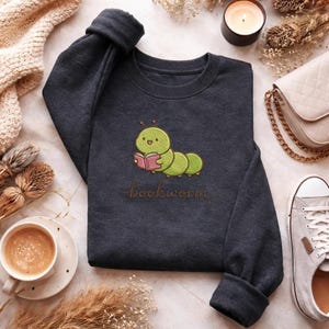 May include: Dark gray sweatshirt featuring a cartoon green caterpillar reading a pink book. The word "bookworm" is written in a cursive font below the illustration. The sweatshirt is made of a soft material.