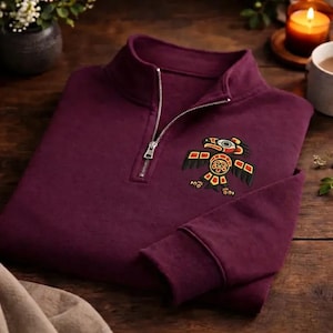 May include: A burgundy quarter-zip sweatshirt with a detailed embroidered Native American-inspired design on the chest. The design features a stylized bird in black, red, and yellow. The sweatshirt has a silver zipper and a soft, comfortable appearance.