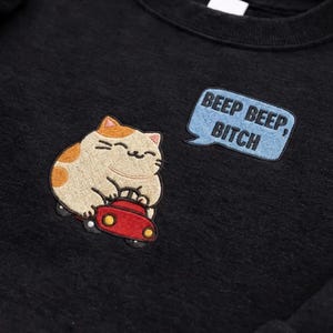 May include: Black sweatshirt with an embroidered design featuring a smiling cat driving a red car. The cat has tan and orange markings. A blue speech bubble with the text "BEEP BEEP, BITCH" is embroidered next to the cat.