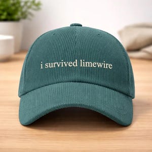 May include: Teal corduroy baseball cap with the phrase "i survived limewire" embroidered in white. The cap has a curved brim and a button on top. A casual accessory.