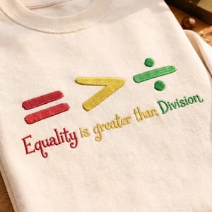 May include: Cream-colored sweatshirt with embroidered text and symbols. The design includes red equal signs, a yellow greater-than symbol, and a green division symbol. The phrase "Equality is greater than Division" is embroidered in a mix of red, yellow, and green.