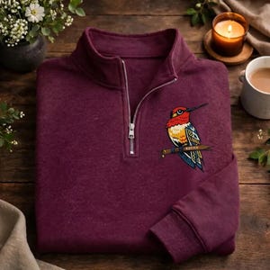 May include: A burgundy quarter-zip sweatshirt with a colorful embroidered hummingbird design on the front. The hummingbird is perched on a branch and has red, yellow, blue, and orange details. The sweatshirt has a silver zipper and a ribbed cuff.