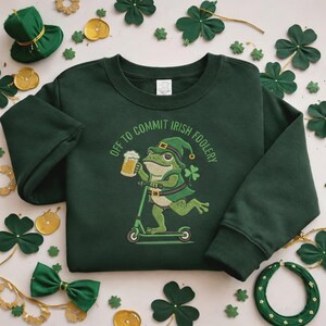 May include: Dark green sweatshirt with the phrase "Off to Commit Irish Foolery" encircling a cartoon frog riding a scooter and holding a beer. The frog wears a green hat and a shamrock. Green shamrocks and a green hat are scattered around the sweatshirt.