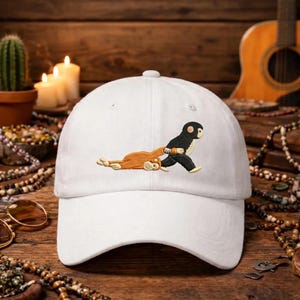 May include: A white baseball cap with an embroidered design. The design features a black and white cartoon character pulling a brown cartoon character. The cap has a curved brim and a button on top. The hat is made of a soft material.