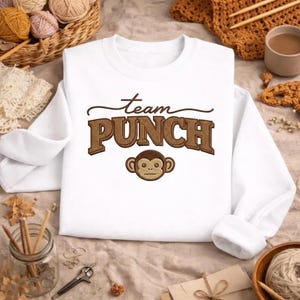 May include: White sweatshirt with the text "Team PUNCH" in brown, featuring a monkey face graphic. The sweatshirt has a crew neck and long sleeves. The design is embroidered on the front.