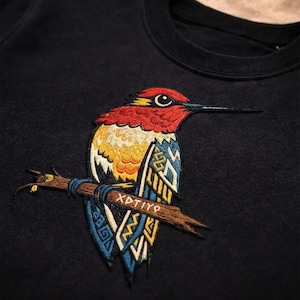 Puede incluir: Sudadera negra con un diseño de pájaro bordado. El pájaro tiene detalles rojos, amarillos y azules, posado en una rama marrón. El texto "XDTiYp" está bordado en la rama. La sudadera está hecha de un material suave.
