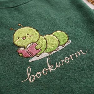May include: Green sweatshirt with an embroidered design of a smiling green caterpillar reading a pink book. The word "bookworm" is embroidered in cursive below the caterpillar.