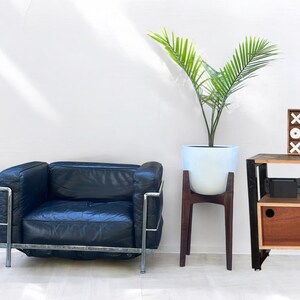 May include: A modern living room scene featuring a black leather armchair with a chrome frame, a white planter with a green plant on a dark wood stand, and a wooden side table with a decorative XOX block.