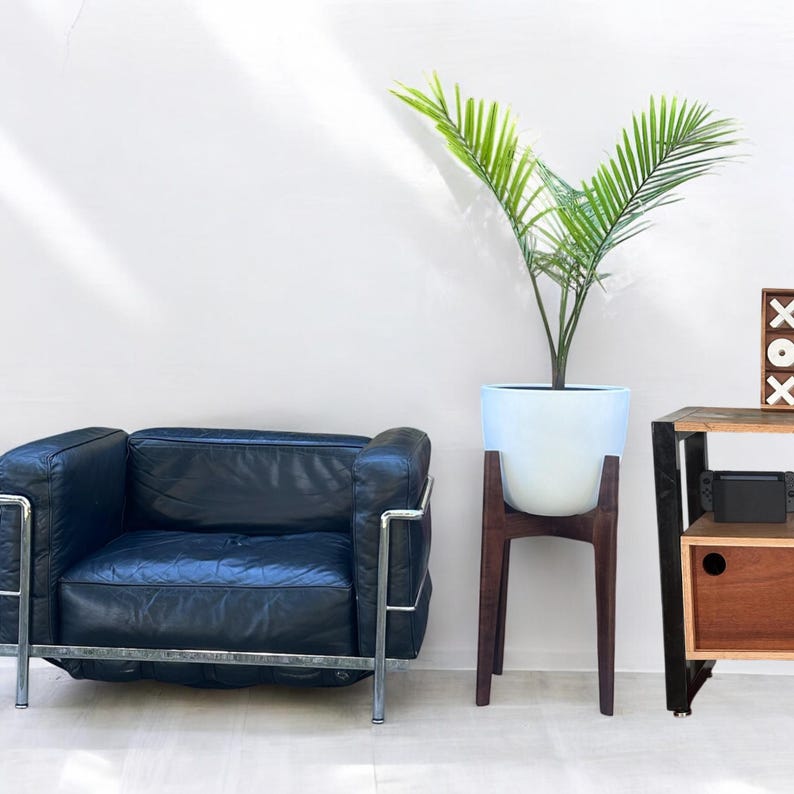 May include: A modern living room scene featuring a black leather armchair with a chrome frame, a white planter with a green plant on a dark wood stand, and a wooden side table with a decorative XOX block.