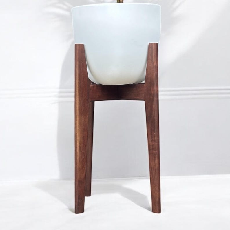 May include: A white ceramic planter sits atop a mid-century modern style wooden plant stand. The stand has a dark brown finish and a minimalist design, with four angled legs. The planter is a simple, rounded shape.